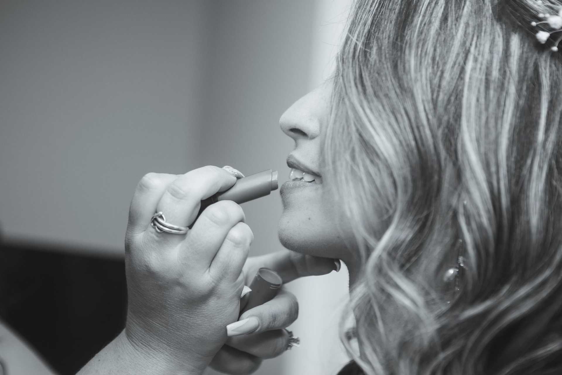 Person applying lipstick to a smiling woman with wavy hair in a close-up shot.