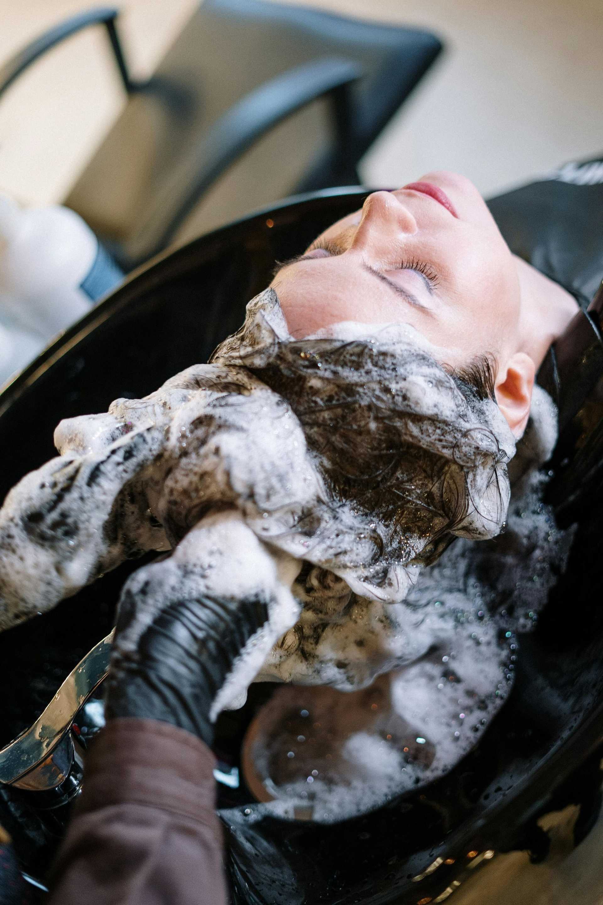 Person having hair washed at a salon, leaning back with eyes closed, hair covered in suds.